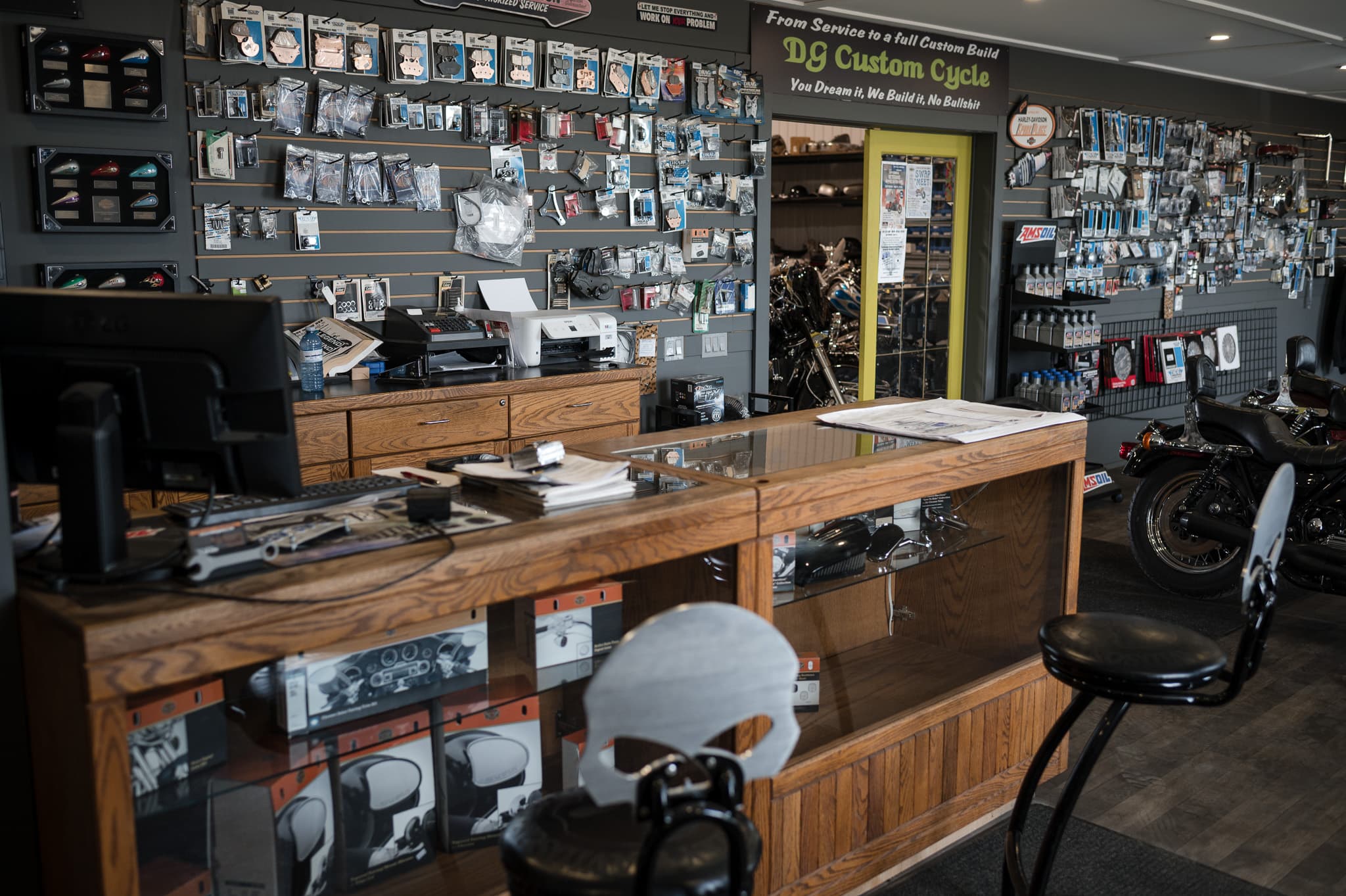 DG Custom Cycle parts counter and showroom, the shop wordmark above the parts wall with a Harley parked alongside