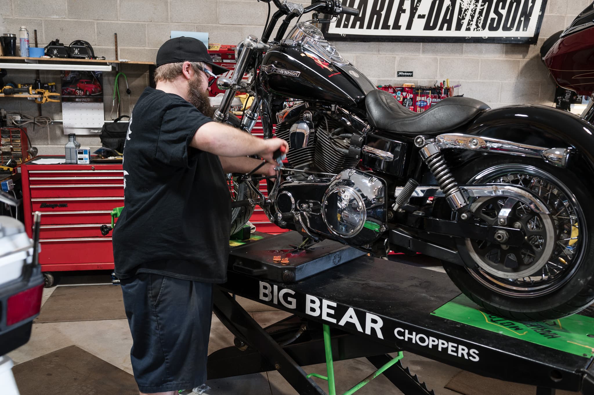 Tech wrenching on a black Dyna up on the Big Bear lift under the shop's Harley-Davidson sign